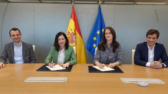Two women sitting at a table signing a document and two men next to them. 