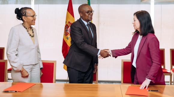 Tres representantes institucionales participan en un acto oficial. Dos de ellos se dan la mano frente a una mesa con documentos, con banderas institucionales al fondo. 